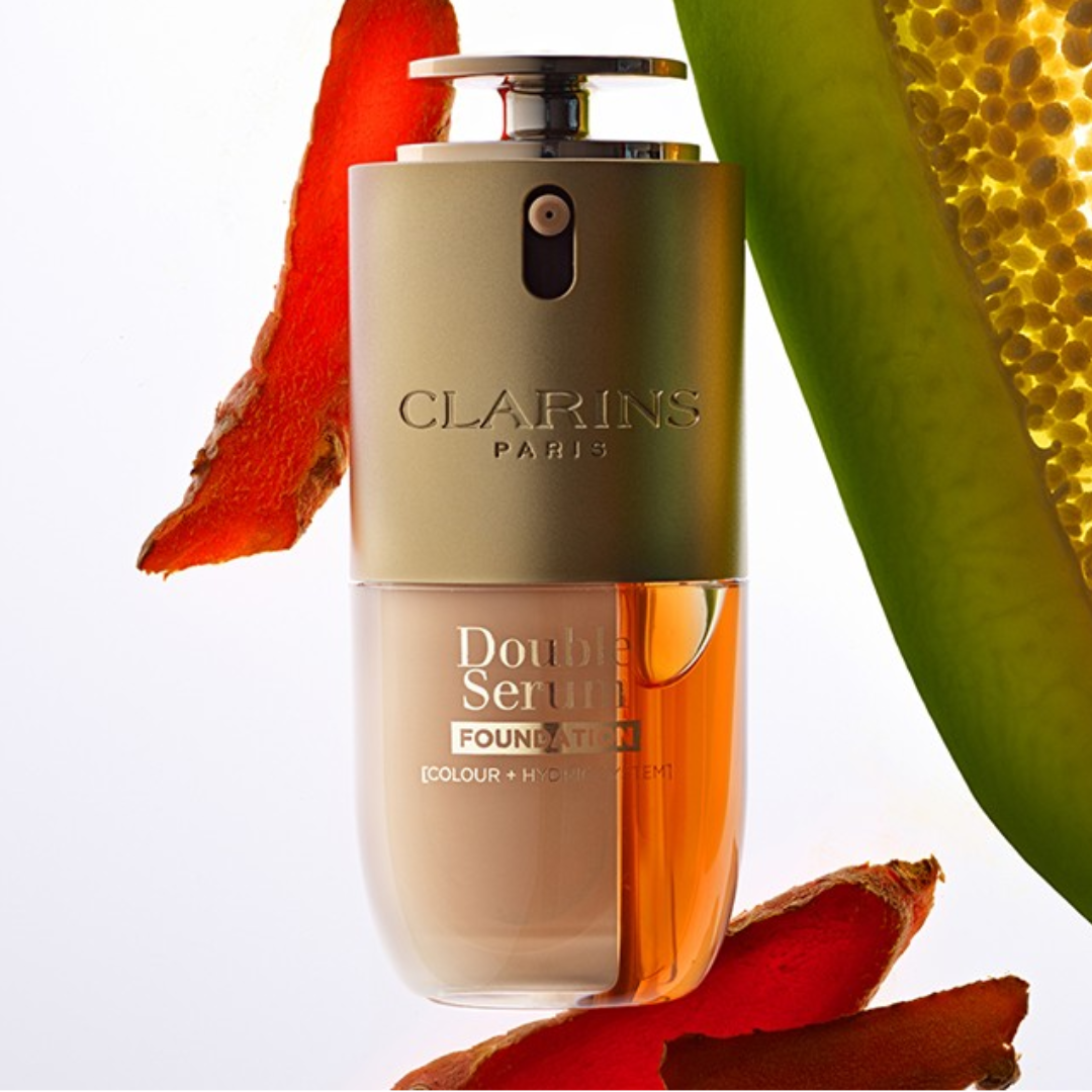 Double Serum Foundation von Clarins mit zweiphasiger Formel in einem Flakon, umgeben von natürlichen Inhaltsstoffen wie Frucht- und Pflanzenextrakten.